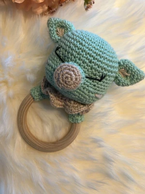 Virkad grön babyskallra med gris - Söt handgjord babyskallra i form av en grön gris med virkade detaljer och ett träringsgrepp. Grisen har stängda ögon, beige nos och öron, samt en liten krage i beige. Perfekt som present till en nyfödd eller babyshower.