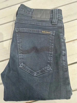 Mörkblå jeans från Nudie Jeans - Snygga mörkblå jeans från Nudie Jeans med klassisk femficksdesign och diskret broderad bakficka. Jeansen har raka ben och är tillverkade i denim med en lätt stretch för extra komfort. Perfekta för en avslappnad och trendig look.