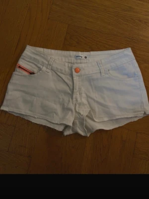 Vita denimshorts med orange knapp - Säljer ett par vita denimshorts med orange knapp och råa kanter. Shortsen har fem fickor, bälteshällor och en liten detaljficka med orange dragkedja. Perfekta för varma dagar och enkel att matcha med olika toppar.