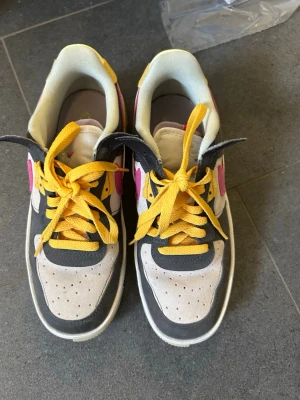 Airforce 1 sneakers - Snygga sneakers i vitt, grått och beige med detaljer i rosa och gult. Skorna har gula skosnören och en klassisk låg siluett. Materialet är en mix av mocka och textil, och designen har perforerade partier framtill för extra ventilation. 