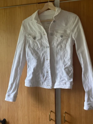 Vit jeansjacka med klassisk passform - Snygg vit jeansjacka med klassisk krage, knäppning framtill och två bröstfickor med lock och knappar. Jackan har långa ärmar och justerbara knappar i midjan för en schysst passform. Perfekt att slänga över en t-shirt för en fräsch look.