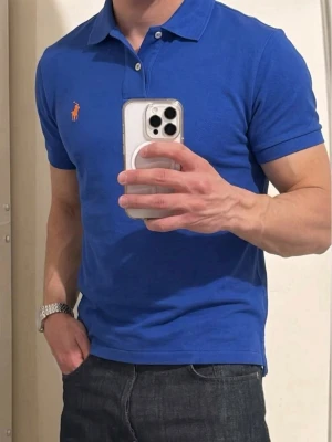 Blå pikétröja från Polo Ralph Lauren - Klassisk blå pikétröja från Polo Ralph Lauren med korta ärmar och krage. Tröjan har den ikoniska broderade loggan i orange på bröstet och stängs med två knappar vid halsen. Perfekt till jeans eller shorts för en clean och sportig look.