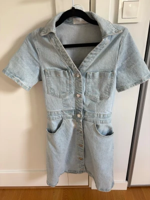 Ljusblå jeansklänning Zara  - Säljer en kort ljusblå jeansklänning från Zara med knappar framtill, krage och fyra fickor. Klänningen har korta ärmar och markerad midja för en snygg passform. 