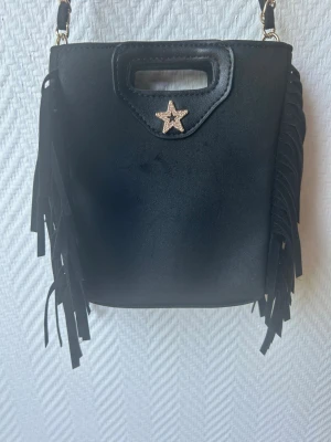 Svart axelväska med fransar och stjärna - Snygg svart axelväska med långa fransar på sidorna och en kedjerem. Väskan har ett dekorativt stjärnformat smycke framtill och ett litet handtag upptill. Materialet ser ut som konstläder och väskan har en cool, trendig look.