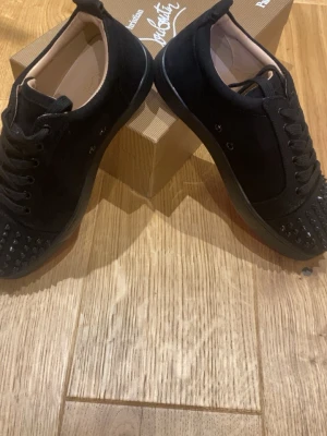Svarta Christian Loubutin med nitar - Svarta sneakers i mocka med coola svarta nitar på tån och svarta skosnören. Snygg och stilren design med låg profil och gummisula. Perfekta för dig som vill sticka ut med en edgy look.