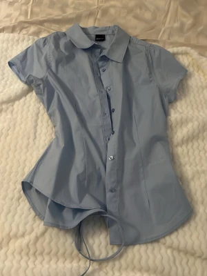 Ljusblå kortärmad skjorta med knyt - Snygg ljusblå skjorta med korta ärmar och klassisk krage. Skjortan har knappar framtill och ett knytband i midjan för en mer markerad siluett. Perfekt till jeans eller kjol för en clean look.