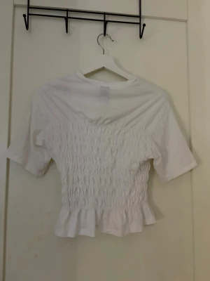 Vit smockad topp med volang - Säljer en söt vit topp med smockad midja och volangkant nedtill. Toppen har korta ärmar och en croppad passform som ger en trendig look. Perfekt att matcha med höga jeans eller kjol.