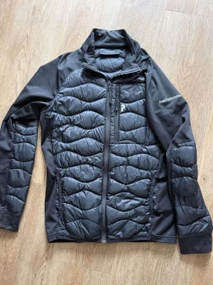 Svart pufferjacka Peak Performance - Snygg svart pufferjacka från Peak Performance med quiltat mönster och hel dragkedja framtill. Jackan har en hög krage, diskret logga på bröstet och ärmar i stretchigt material för extra komfort. Perfekt för kalla vinterdagar.