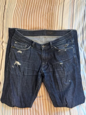 Mörkblå slitna jeans från Tiger of Sweden - Säljer ett par mörkblå jeans med raka ben och slitna detaljer på framsidan. Det är egen designade så hålen har gjorts själv. Extremt snygga och väldigt sköna!