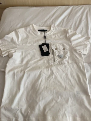 Vit t-shirt från Prada med ficka - Säljer en stilren vit t-shirt från Prada i storlek S. T-shirten har en unik bröstficka med dragkedja och metallknappar samt Pradas ikoniska metallogo. Tillverkad i mjuk bomull och har klassisk passform med rund hals och korta ärmar.