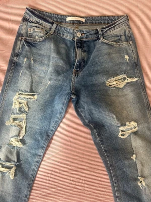 Blå slitna jeans med hål - Säljer ett par blå jeans med flera slitningar och hål på både fram- och baksida. Jeansen har en klassisk femficksmodell, normal midja och smal passform. Materialet är denim i bomull och de har en cool, avslappnad vibe.