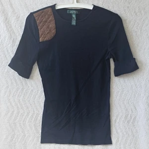 Mörkblå ribbad topp från Ralph Lauren - Snygg mörkblå ribbad topp från Lauren Ralph Lauren med korta ärmar och en brun quiltad detalj på axeln. Tillverkad i mjuk bomull och polyester. Perfekt för dig som gillar klassisk men unik stil.