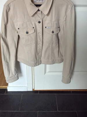 Beige jeansjacka med fickor - Stilren beige jeansjacka med klassisk krage och två bröstfickor med knappar. Jackan har långa ärmar, knappstängning framtill och detaljerade sömmar. Materialet är stretch denim, väldigt skön och modellen är rak och kortare i passformen.
