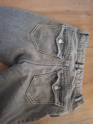 Grå jeans från Lindex, modell Freja - Snygga grå jeans från Lindex, modell Freja. Jeansen har raka ben, klassiska fem fickor och silverfärgade knappar. Tillverkade i bomullsmix med en tvättad look och kontrastsömmar. Perfekta för en avslappnad och trendig stil.