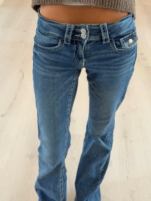 Blå jeans med låg midja - Snygga blå bootcut jeans med låg midja och fickor. Jeansen har dubbla knappar i midjan och ljusa slitningar framtill för en trendig look. Perfekta för dig som gillar en avslappnad men ändå stilren stil. Storlek 152 TALL.