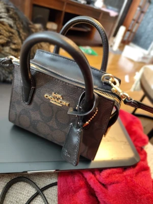 Mörkbrun miniväska från Coach - Snygg mörkbrun minihandväska från Coach med klassiskt monogrammönster och guldfärgade detaljer. Väskan har två handtag, avtagbar axelrem och dragkedja upptill. Tillverkad i tåligt läder med Coach-logga och vagn i metall framtill.