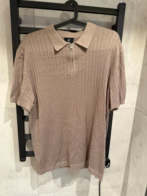 Beige ribbad polotopp med dragkedja - Säljer en beige ribbad polotopp från H&M med korta ärmar och en snygg dragkedja framtill. Toppen har en klassisk krage och är tillverkad i mjukt material som sitter skönt på kroppen. Perfekt för dig som gillar stilrena och enkla plagg.