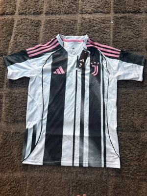 Juventus matchtröja Slim fit - Juventus officiella matchtröja från Adidas i storlek S men sitter som M. Tröjan har svartvita ränder med rosa detaljer på axlar och loggor. Tillverkad i ett lätt och ventilerande material, perfekt för fotboll eller träning. Slim fit-modell med klubbmärke och tre stjärnor på bröstet.