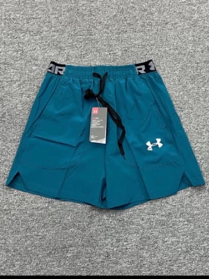 Blå träningsshorts från Under Armour - Snygga blå träningsshorts från Under Armour med elastisk midja, svart snörning och logga på vänster ben. Shortsen har praktiska fickor och är perfekta för gymmet eller löprundan.