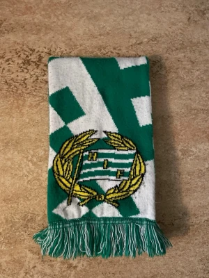 Grönvit Hammarby halsduk med tofsar - Säljer en grön och vit halsduk med Hammarby IF:s klassiska klubbmärke i gult och grönt. Halsduken har rutigt mönster och fransar i ändarna. Perfekt för dig som vill visa ditt stöd på matchen eller bara gillar snygga accessoarer.