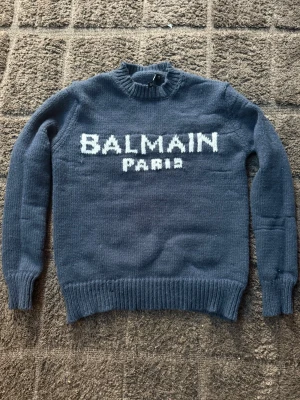 Mörkblå stickad tröja Balmain Paris - Mörkblå stickad tröja från Balmain Paris med vit logotyp framtill. Tröjan har rund halsringning, ribbstickade muddar och nederkant. Perfekt för dig som gillar exklusiva och trendiga plagg med tydlig logga.