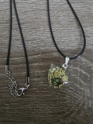 Halsband med svart band och blomma - Unikt halsband med svart läderliknande band och ett droppformat hänge i transparent glas. Hänget innehåller små gula torkade blommor och en fjäril, vilket ger en naturlig och bohemisk känsla. Silverfärgat spänne och justerbar kedja.