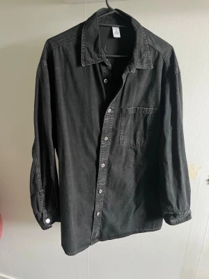 Svart långärmad skjorta i tunnare jeans - Stilren svart skjorta med klassisk krage och bröstficka. Skjortan har långa ärmar och knäppning framtill.  Perfekt att bära öppen över t-shirt eller stängd.