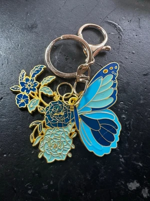 Nyckelring med blå fjäril och blommor - Snygg väsksmycke/nyckelring i metall med emaljdetaljer, föreställande en blå fjäril och blommor i olika blå nyanser med guldkant. Perfekt att hänga på väskan eller nyckelknippan för en färgglad touch.