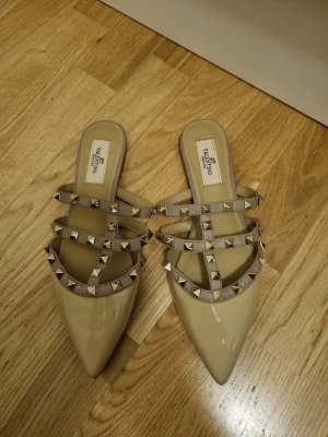 Beige sandaler med nitar från Valentino - Säljer ett par snygga beige sandaler från Valentino med spetsig tå och flera remmar dekorerade med guldfärgade nitar. Skorna har öppen häl och är tillverkade i glansigt material som ger en lyxig känsla. Perfekta för dig som vill sticka ut med din stil.