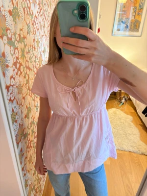 Ljusrosa blus med rosett från Boomerang - Supersöt ljusrosa blus från Boomerang med korta ärmar och knyt i ringningen. Blusen har en avslappnad passform och lätt rynkad midja som ger en feminin siluett. Perfekt till jeans eller kjol för en somrig look. 