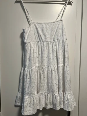 Vit ärmlös sommarklänning med volang - Söt vit klänning med tunna axelband och volangkant. Klänningen har broderade detaljer och är ärmlös, perfekt för varma dagar. Lätt och luftig modell som ger en avslappnad känsla.