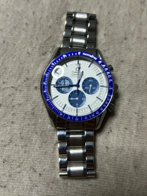 Omega Speedmaster blå/silver klocka - Snygg Omega Speedmaster rep kronograf med blå och vit urtavla, blå tachymeter-ring och robust länkarmband i silverfärgat stål. Baksidan har en cool mån- och jorddetalj. Perfekt för dig som gillar klassisk men sportig stil.