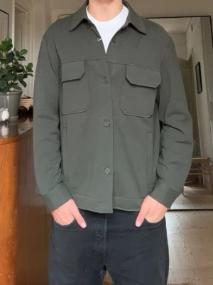 Mörkgrön overshirt med fickor - Stilren mörkgrön overshirt med två stora bröstfickor och knäppning framtill. Skjortkrage och raka ärmar ger en clean look. Tillverkad i ett mjukt, lite tjockare tyg som passar perfekt som lager. Enkel att matcha med olika outfits.