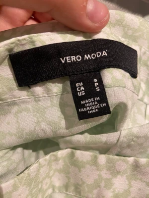 Blommig omlottkjol från Vero Moda - Söt ljusgrön omlottkjol från Vero Moda med vitt blommönster och volangkant nedtill. Kjolen har knytband i midjan och är perfekt för vår och sommar. Lätt och luftig känsla. Nästa aldrig använd