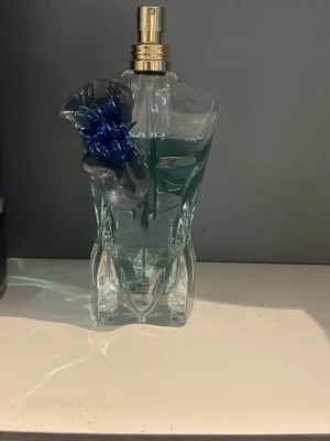 Jean Paul Gaultier Le Beau Flower Edition - Unik parfymflaska formad som en manlig torso med blå och guld detaljer. Flaskan har en dekorativ blå blomma på axeln och innehåller en blågrön vätska. Kommer i en snygg cylinderförpackning med färgstarkt blommotiv och guldfärgad text. Har inte tatt så många spray som ni ser så det är fortfarande mycket kvar.