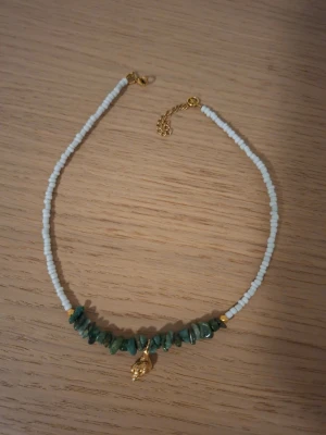 Halsband med vita pärlor och grön sten - Snyggt halsband med små vita pärlor och gröna stenbitar framtill. I mitten hänger ett litet guldfärgat snäckskal som detalj. Halsbandet har guldfärgat lås och justerbar kedja. Perfekt accessoar för att lyfta din outfit.