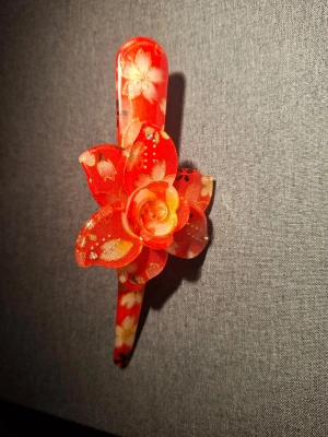Röd hårklämma med blomma - Snygg hårklämma i metall med röd plastdekoration och en stor, detaljerad blomma i tyg och plast. Klämman har blommönster i vitt och guld på både blomman och själva klämman, vilket ger en färgstark och unik look.