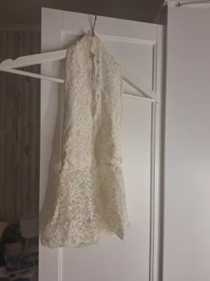 Vit spetsig halterneck topp - Säljer en vit halterneck topp i genombruten spets med blommönster. Toppen har en figurnära passform och öppen rygg, perfekt för att styla lager på lager eller till ett par höga jeans. Materialet är mjuk och stretchig spets.