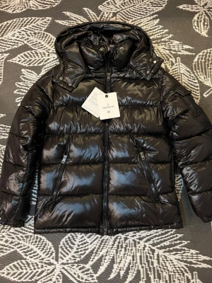 Svart pufferjacka från Moncler - Snygg svart pufferjacka från Moncler med glansig finish, huva och dragkedjor på fickorna. Jackan har en klassisk quiltad design och är perfekt för kalla vinterdagar. Märkeslogga på ärmen och justerbar huva för extra komfort.