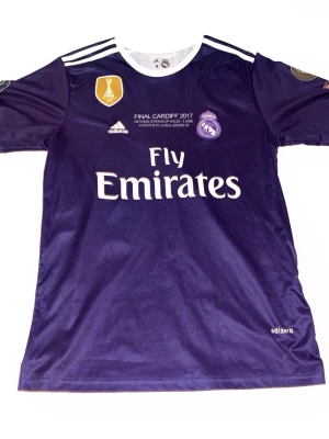 Lila Real Madrid matchtröja Adidas - Säljer en lila Real Madrid matchtröja från Adidas med vita detaljer och tryck. Tröjan har tryck för Champions League-finalen 2017 i Cardiff, klubbmärke, Adidas-logga och Fly Emirates sponsortryck. Tillverkad i lätt och ventilerande material.