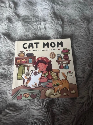 Cat Mom målarbok - En söt och rolig målarbok med titeln 'Cat Mom'. Omslaget visar en tecknad kvinna och två katter i ett kök. Boken har färgglada illustrationer och flera sidor med kattmotiv att färglägga. Perfekt för kattälskare som gillar att vara kreativa.