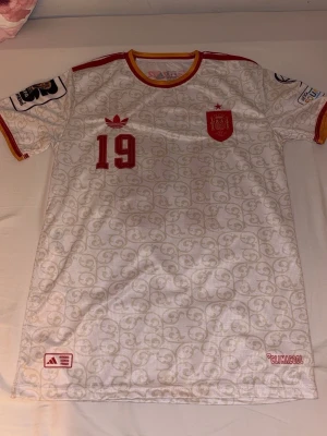Vit Spanien fotbollströja Adidas - Säljer en vit Spanien fotbollströja från Adidas med röd och gul krage, röda detaljer och nummer 19 på bröstet. Tröjan har ett subtilt mönster över hela tyget, broderad landslagssköld och patchar på ärmarna. Perfekt för match eller träning.