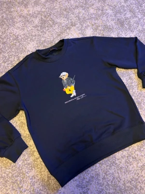 Mörkblå Ralph lauren  - Mörkblå sweatshirt från Polo Ralph Lauren med ikoniskt björntryck på bröstet. Tröjan har rund hals, ribbade muddar och avslut. Perfekt för dig som gillar streetwear och klassisk stil.