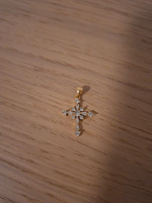 Guldfärgat korshalsband med stenar - Snyggt halsbandshänge i form av ett kors, guldfärgat med gnistrande klara stenar som dekoration. Hänget har en elegant och klassisk design som passar till många olika stilar. Perfekt för dig som gillar detaljer och vill ha något som sticker ut. Doppat i 18k guld.