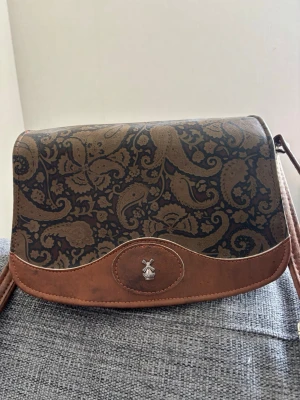 Brun axelväska med paisleymönster - Snygg brun axelväska med paisleymönster i mörkbrunt och beige tillverkad på 90-talet. Väskan har ett lock med dekorativt emblem framtill och justerbar axelrem. Materialet ser ut att vara konstläder och textil, perfekt för dig som gillar vintagevibbar och unika detaljer. Mått 24x18x14 cm 
