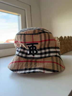 Beige bucket hat med rutigt mönster - Säljer en beige bucket hat med klassiskt rutigt mönster i svart, vitt och rött. Hatten har ett broderat svart TB-emblem framtill och är tillverkad i ett mjukt tyg, perfekt för en trendig och avslappnad stil.