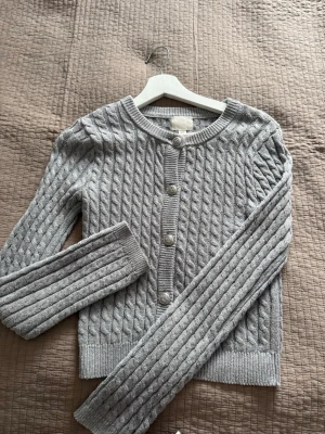 Grå kabelstickad kofta från H&M - Snygg grå kabelstickad kofta från H&M med rund halsringning och dekorativa silverfärgade knappar framtill. Koftan har långa ärmar och ribbade muddar vid ärmslut och nederkant. Perfekt att slänga på sig när det är lite kyligt ute.