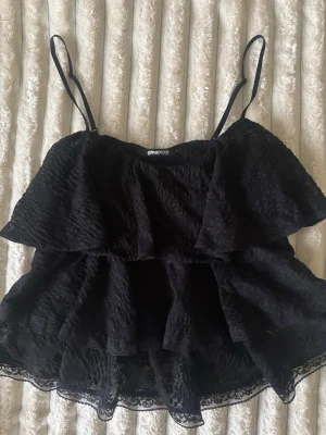 Svart volangtop från Gina Tricot - Säljer en svart linnetopp från Gina Tricot med tunna axelband och flera lager volanger. Toppen har spetsdetaljer längst ner och är perfekt för dig som gillar en luftig och trendig look. Materialet känns lätt och har en snygg struktur.