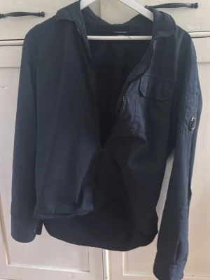 Mörkblå overshirt med dragkedja - Snygg mörkblå overshirt från c.p company med dragkedja framtill och bröstficka. Långärmad modell med klassisk krage och en detaljficka på ärmen. Perfekt att slänga över en t-shirt för en avslappnad look.