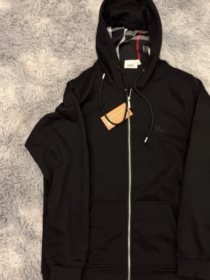 Svart hoodie med dragkedja Burberry - Snygg svart hoodie från Burberry med dragkedja framtill och klassiskt rutigt foder i huvan. Justerbara snören i huvan och två fickor framtill. Diskret Burberry-logga broderad på bröstet. Perfekt för en stilren och avslappnad look.